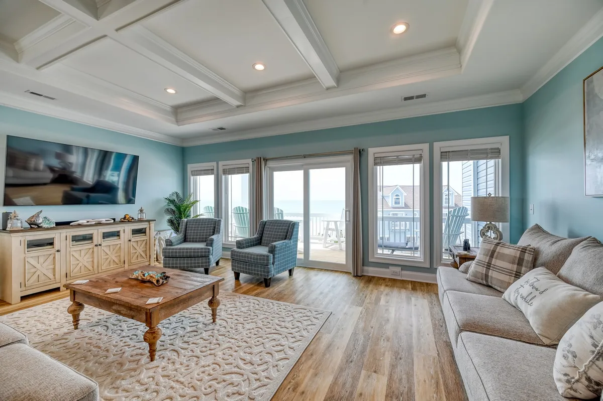 A bright, inviting living room with a large TV on the wall, wooden coffee table, comfortable chairs, and expansive windows framing ocean views. The room blends relaxation and gathering spaces with direct visual access to the water outside.