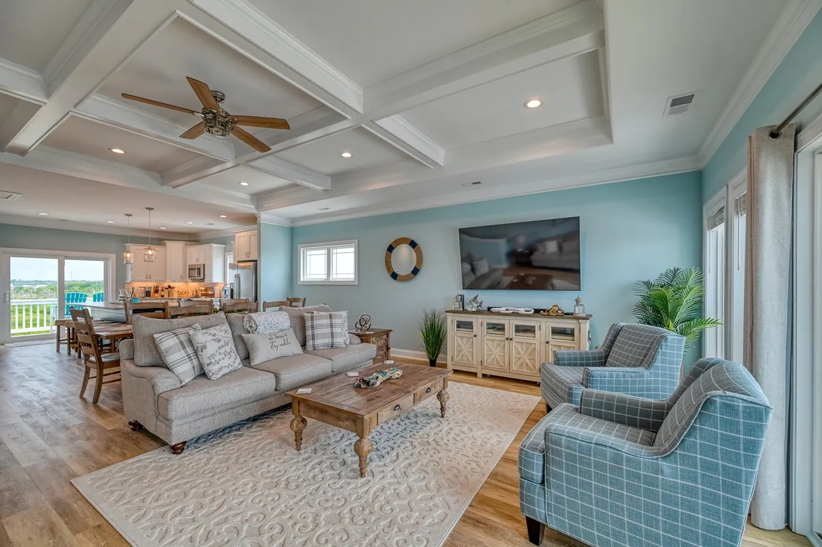 A spacious, sunlit living room with plush sofas arranged around a coffee table, an extra-large television mounted for movie nights, and a dining area in the background. Sliding glass doors lead out to the oceanfront porch, inviting natural light and providing glimpses of the view.