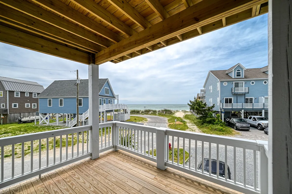 An inviting wooden porch overlooks blue beach houses and a wide beach view. This image evokes the experience of greeting the sunrise or relaxing outdoors with direct lines of sight to the sand and surf.