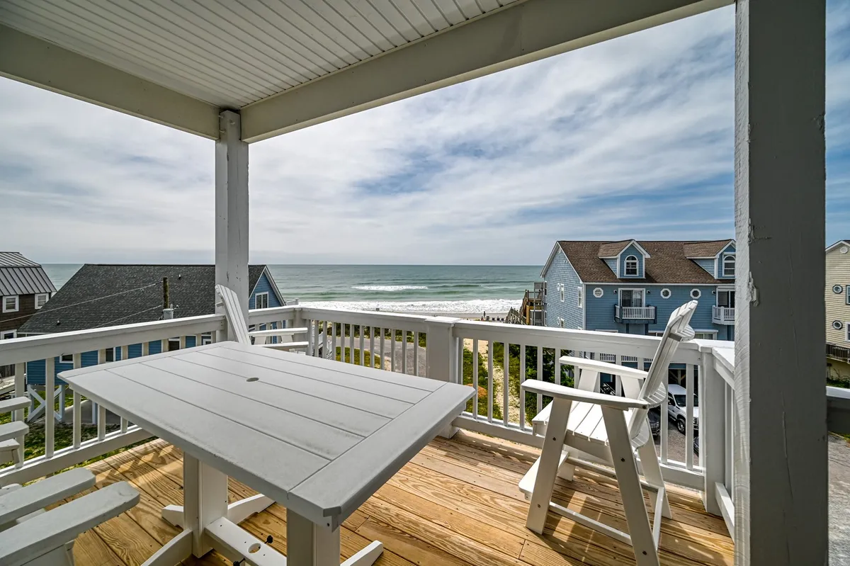 The image showcases a tranquil outdoor dining area set with white chairs and a table, all overlooking sweeping, unobstructed views of the Atlantic Ocean. The deck is bathed in morning sunlight, with the sandy path to the beach visible below, evoking a seamless indoor-outdoor lifestyle and the promise of ocean breezes and starlit evenings.