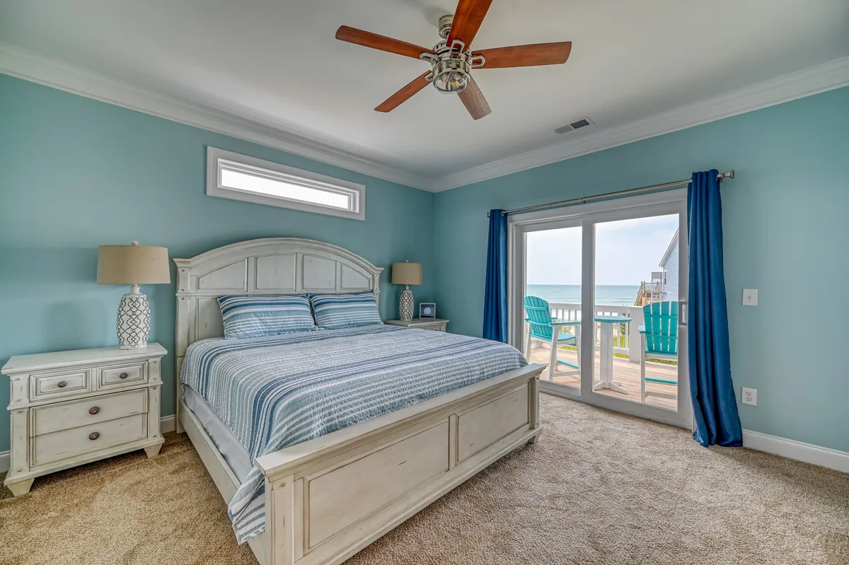A tranquil bedroom with a bed featuring striped bedding, double doors that open to an outdoor deck, and a decorative lamp nearby. Natural light pours in, offering a seamless transition between indoor comfort and outdoor beauty.