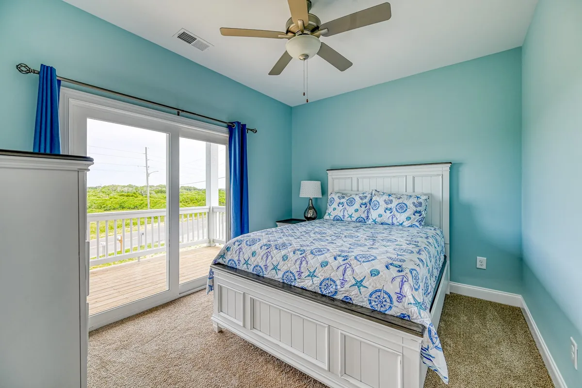 This image features a serene bedroom with blue and white bedding, large windows, and sliding doors that open to a private deck, offering an inviting retreat with tranquil ocean views. The bright and airy design emphasizes comfort and relaxation, making it ideal for a primary suite.