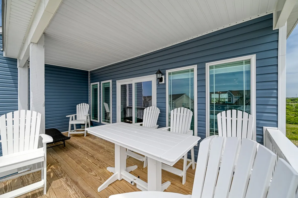 A welcoming outdoor space featuring white Adirondack chairs and a wooden table set against the blue siding of the house. The area is designed for socializing and outdoor dining, with the setting sun casting a warm glow, suggesting a perfect environment for group gatherings and sunset dinners.