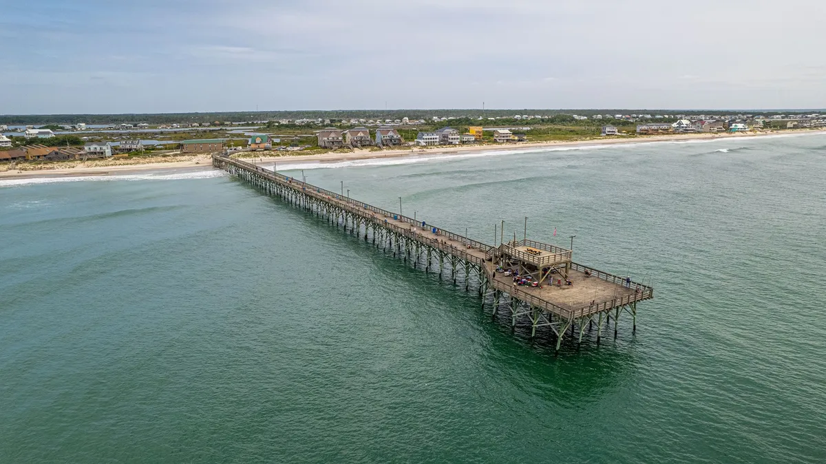 A scenic vista of a pier stretching out into the ocean with the beach in the foreground. The scene evokes a sense of exploration, coastal ambience, and group activities like walks, fishing, or simply soaking in a sunset together.
