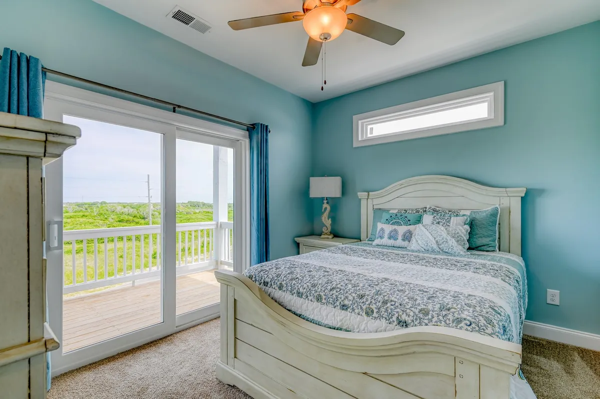 A serene bedroom featuring a bed with patterned bedding, generous windows that provide views of the outdoors, and a cozy nightstand with a lamp. The scene suggests peaceful, private mornings with ample natural light and the potential for ocean vistas.