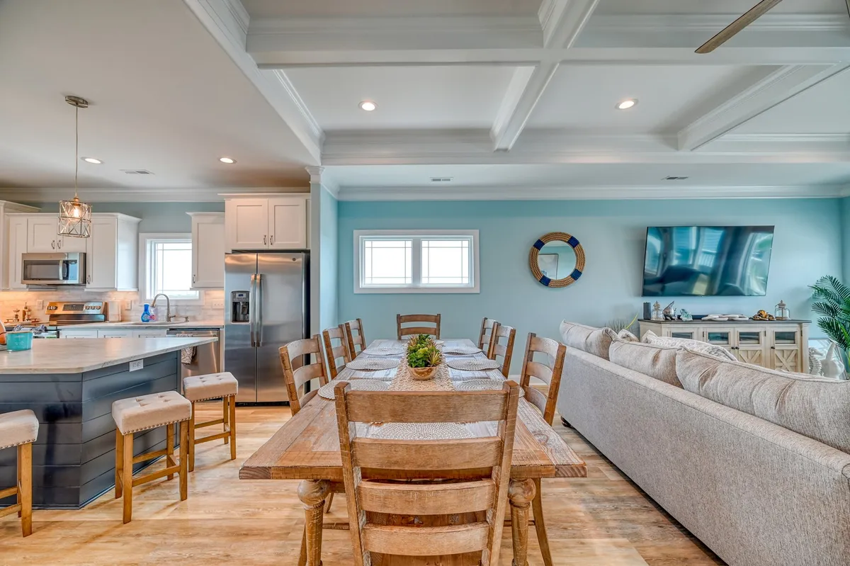 An inviting open-concept space featuring a long French Provincial dining table, a kitchen island with seating, a comfortable sofa, and a television. Decorative touches and natural light create a warm, communal atmosphere ideal for shared meals and celebrations.