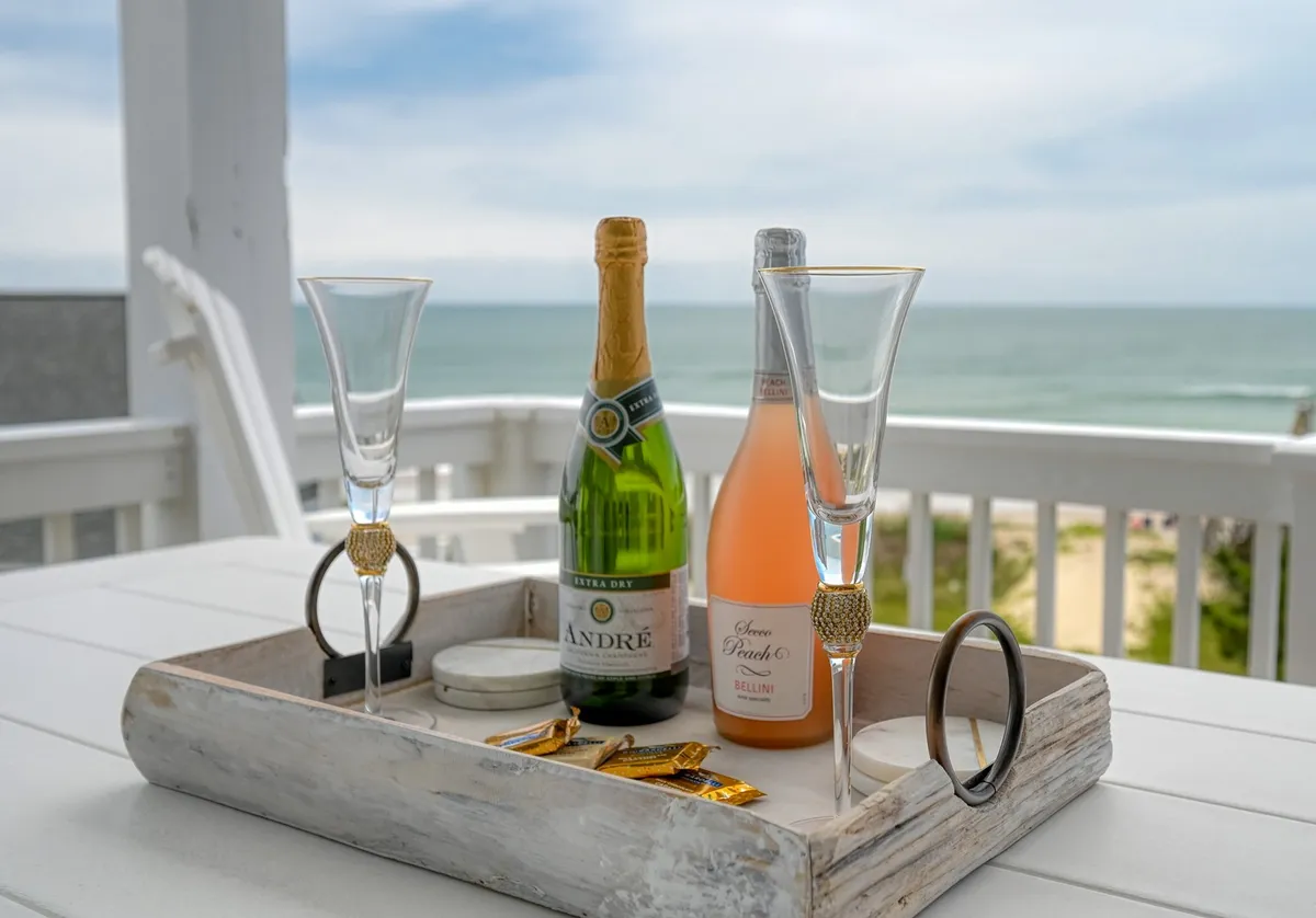 A decorative tray with champagne bottles and flutes sits atop an outdoor surface, suggesting celebration and relaxation. The background hints at an outdoor setting, likely a deck or porch, with the soft glow of sunset reflected in the glass.