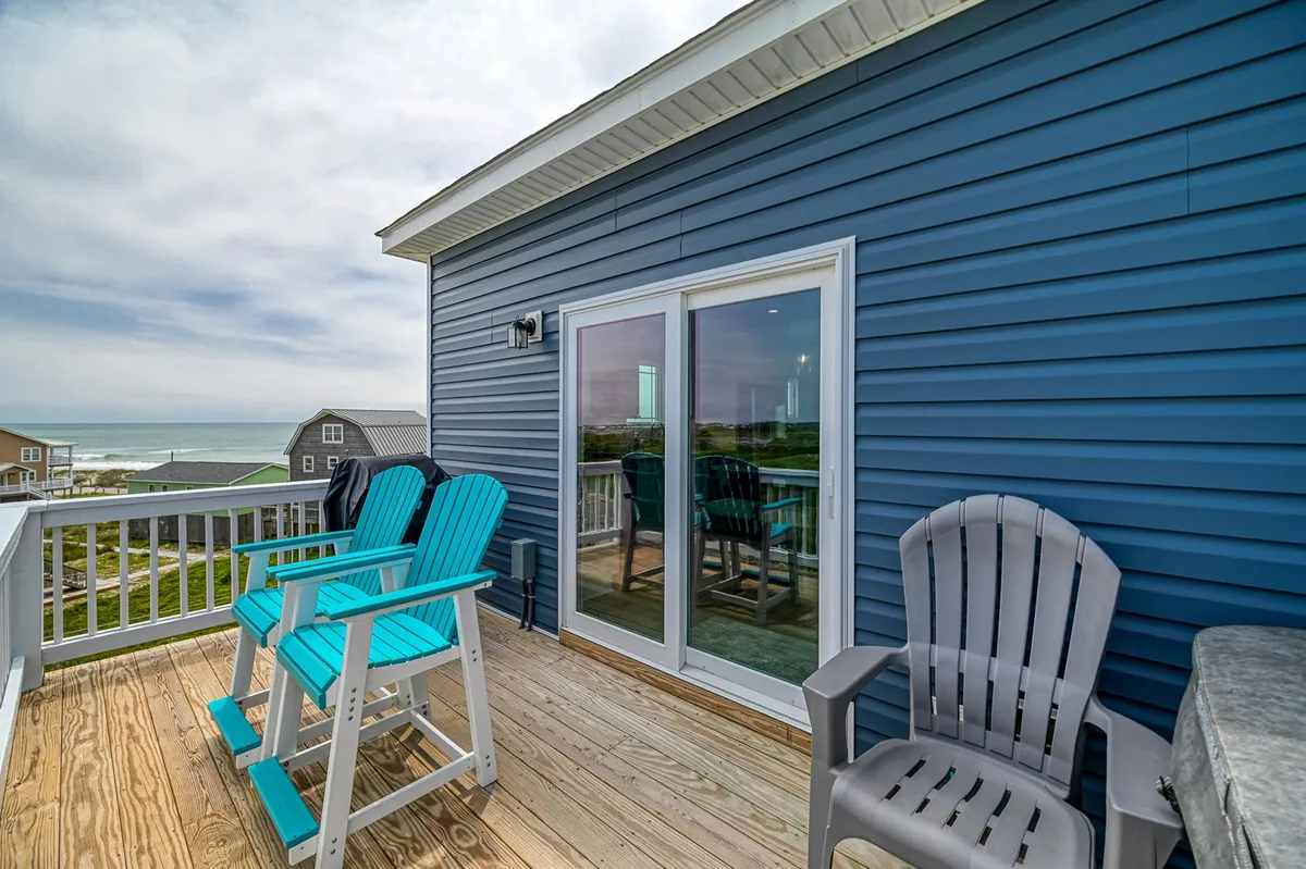 A cozy group of turquoise and gray chairs set on a wooden deck, overlooking the ocean. The sliding glass door suggests immediate indoor-outdoor flow, while the relaxed arrangement encourages socializing or quiet contemplation as the sun sets over the waves.