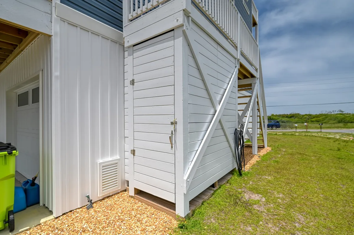 An exterior image showing a landscaped grassy area, a storage shed, and a staircase leading up. It feels casual and relaxed, suggesting ample space to store beach gear and easily manage coming and going from the property.