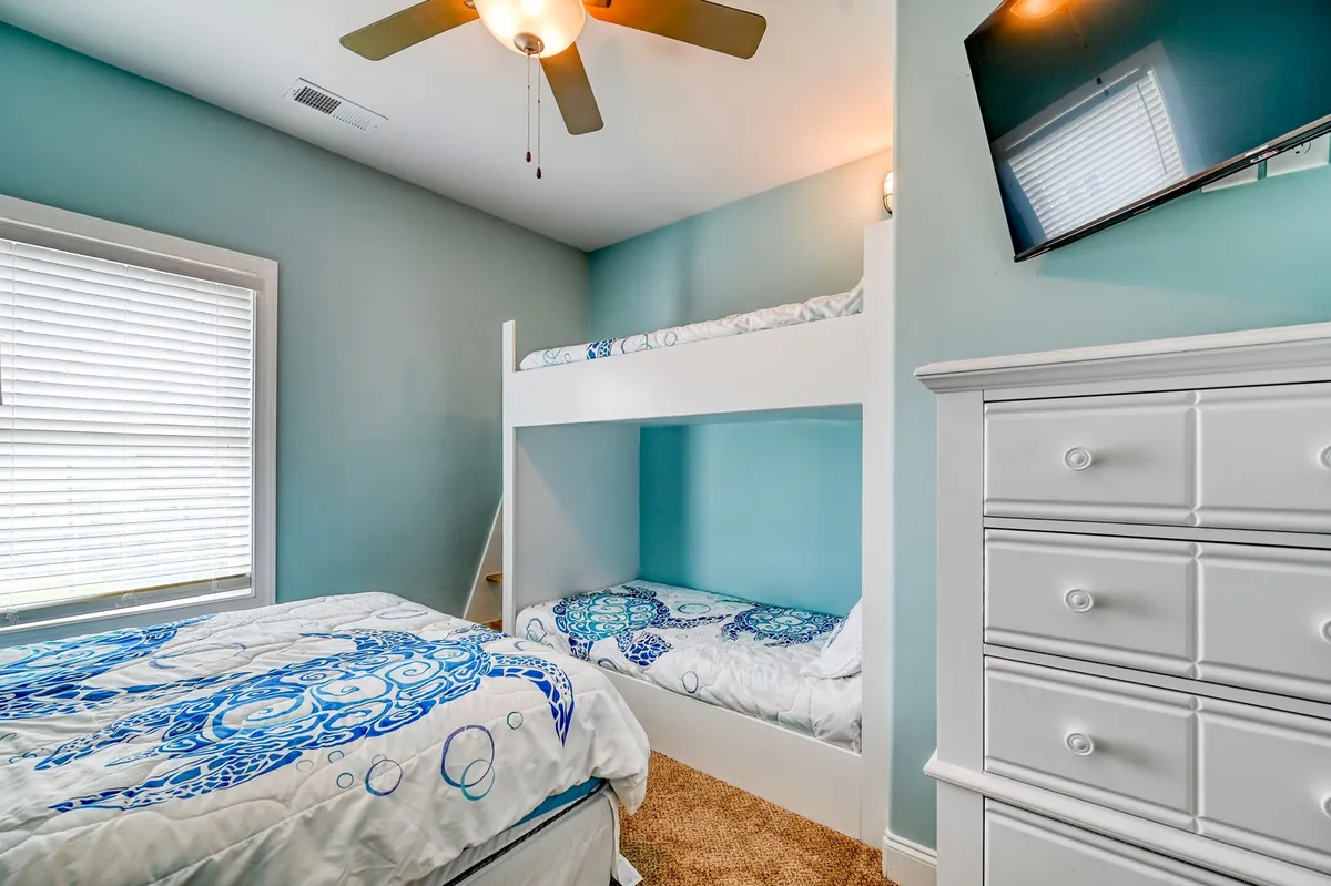A playful and cozy interior bedroom featuring a bunk bed and a regular bed, both adorned with decorative bedding, a dresser, a ceiling fan, and a window with blinds. The atmosphere is inviting, suggesting a family-friendly space where guests feel at home.